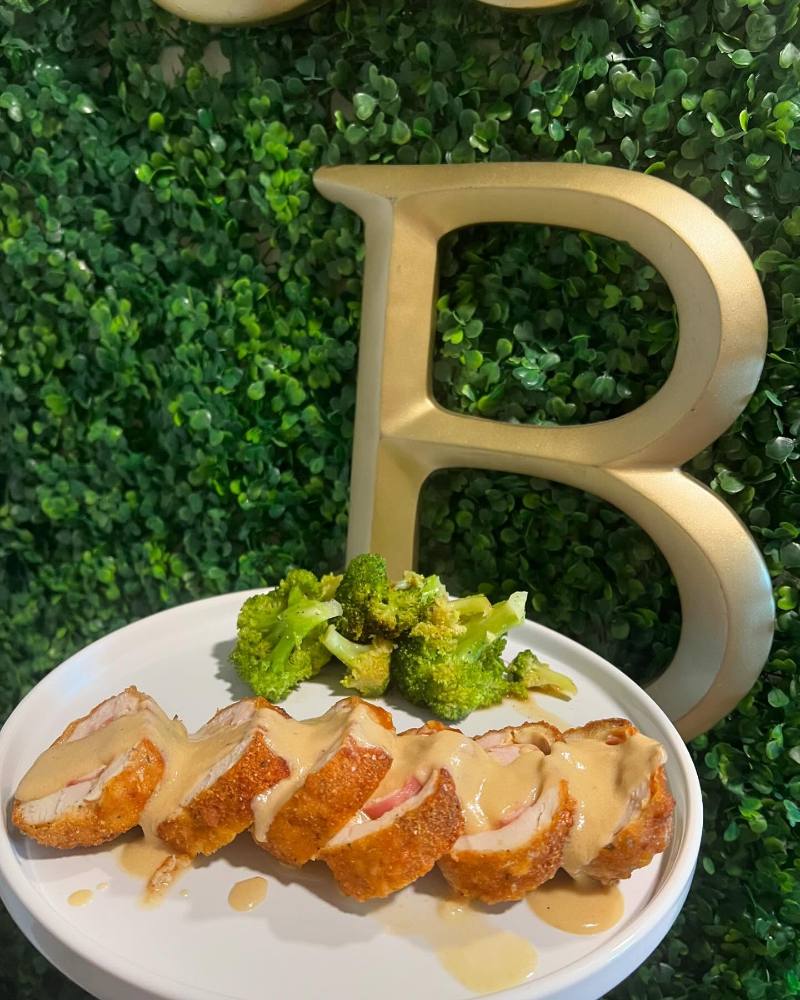 Sliced breaded chicken cordon bleu drizzled with creamy sauce and served with broccoli on a white plate against a green hedge backdrop with a large gold letter B.