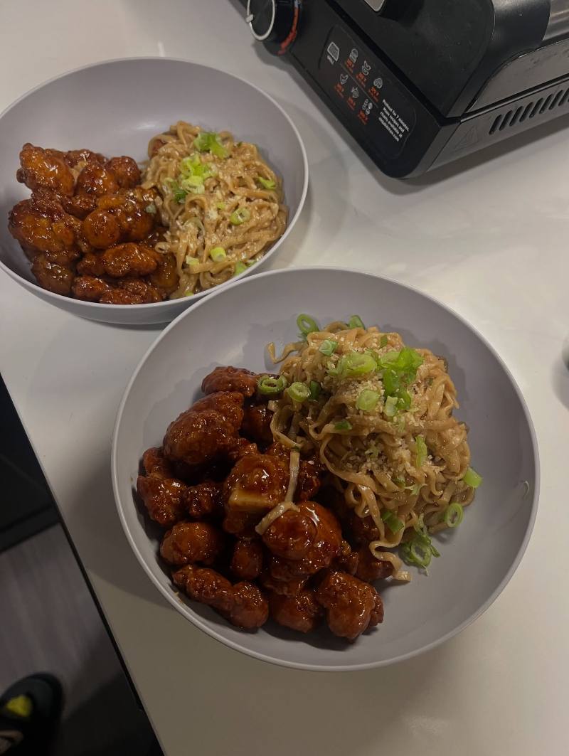 Two gray bowls of glossy sauced chicken with noodles garnished with scallions and sesame seeds sit on a light kitchen countertop beside a black appliance.