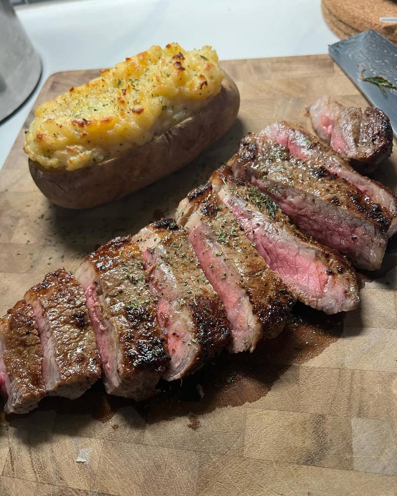 Sliced medium-rare steak with herbs and a golden twice-baked potato are served on a wooden cutting board.