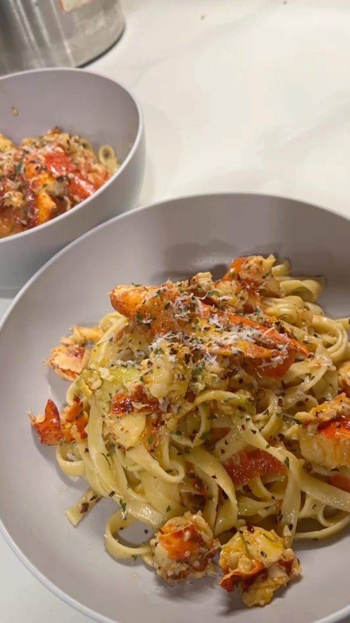 Two gray bowls of lobster pasta with flat noodles, chunks of lobster, herbs, red pepper flakes, and grated cheese on a light countertop.
