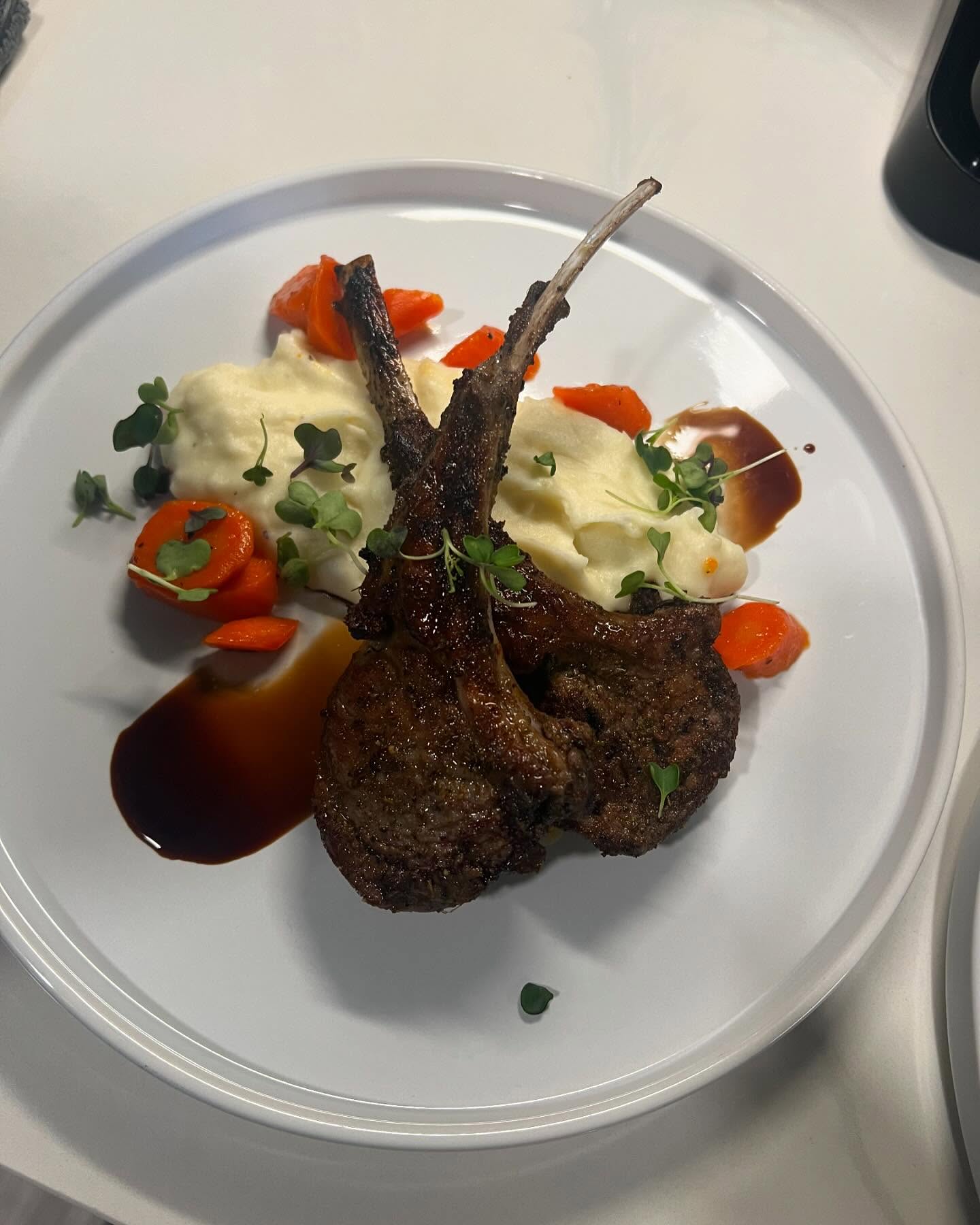 Seared frenched lamb chops plated over creamy mashed potatoes with glazed carrots, dark jus, and microgreens on a white plate set on a light countertop.
