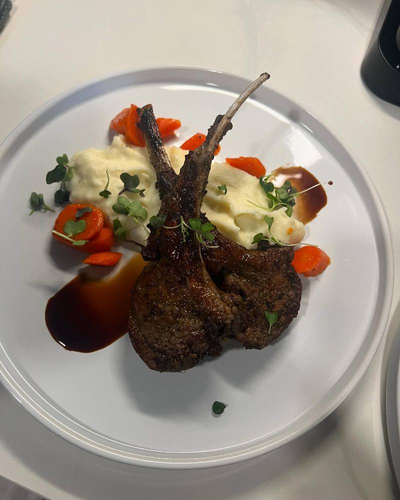 Seared frenched lamb chops plated over creamy mashed potatoes with glazed carrots, dark jus, and microgreens on a white plate set on a light countertop.