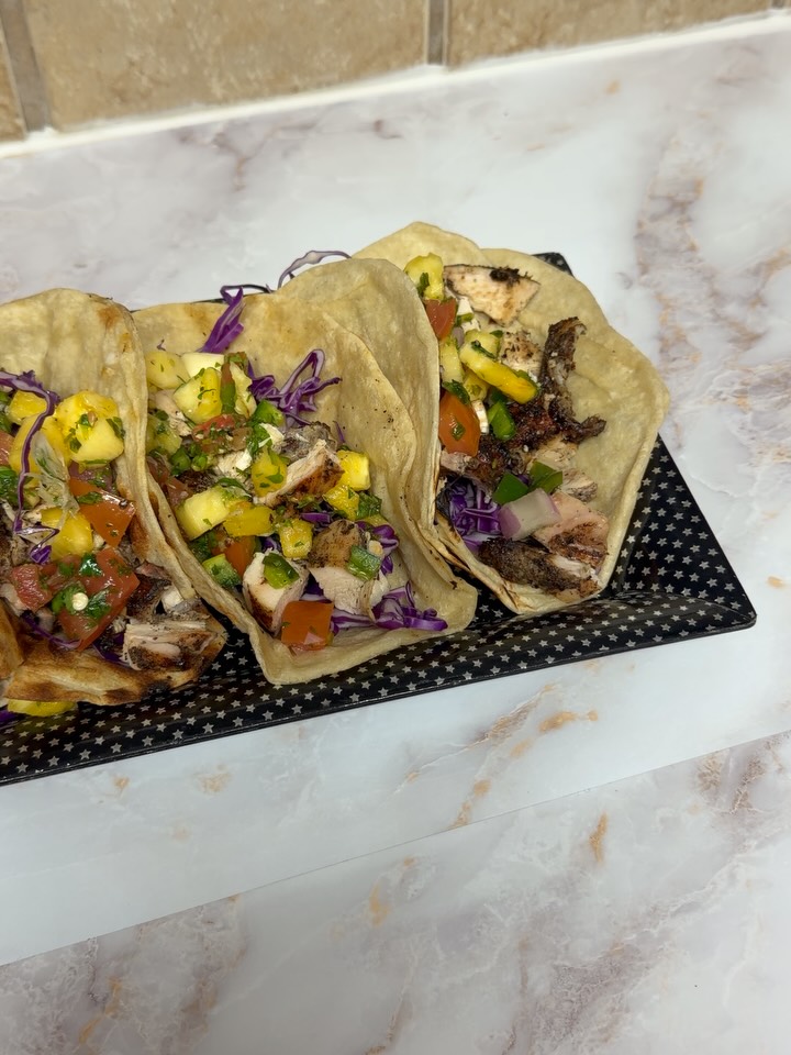 Three grilled chicken tacos with purple cabbage and mango-tomato salsa are served on a black star-patterned rectangular plate on a marble countertop.