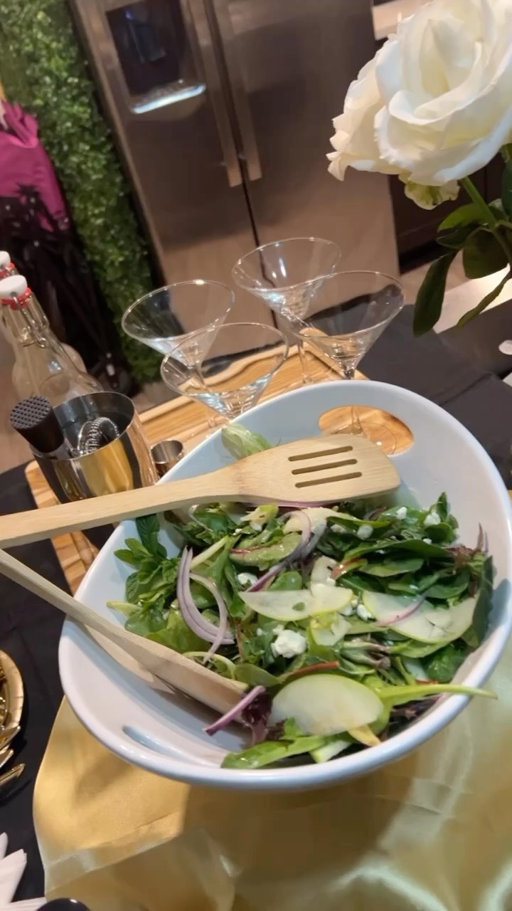 Apple, feta, mint, and red onion salad in a white serving bowl with wooden utensils, set on a gold tablecloth beside martini glasses and a white rose.