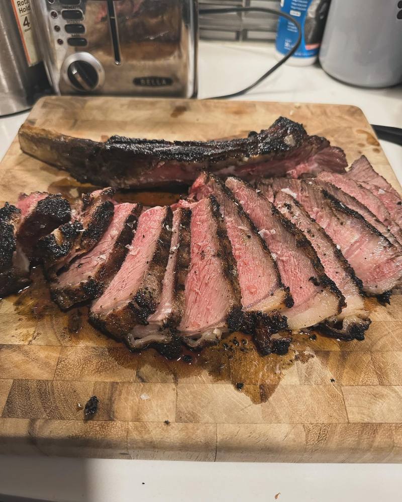 Sliced medium-rare tomahawk ribeye with a charred crust and coarse salt rests beside the bone on a wooden cutting board in a kitchen.