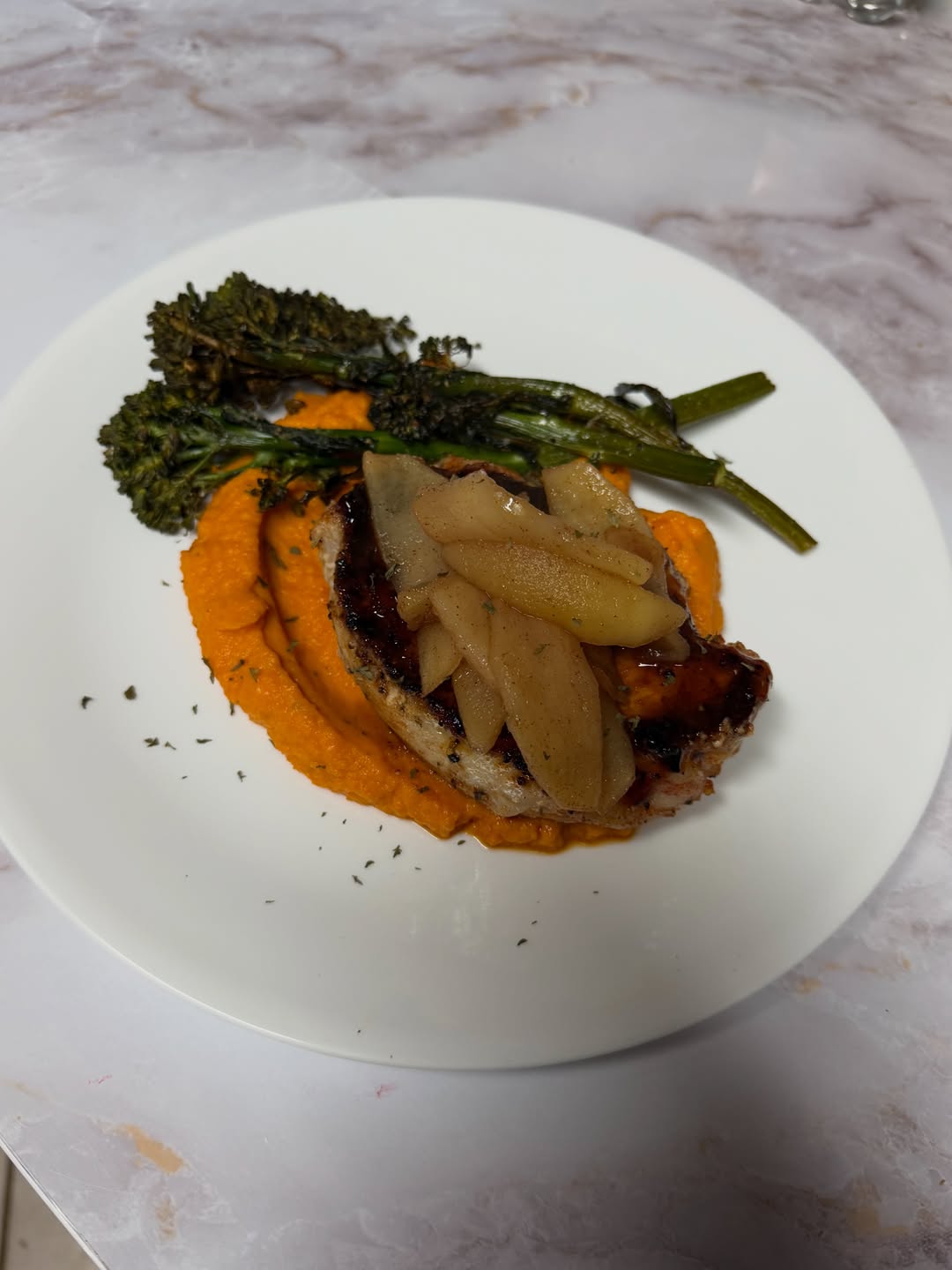 Seared chop topped with sautéed apple slices, served over bright orange purée with charred broccolini on a white plate on a marble countertop.