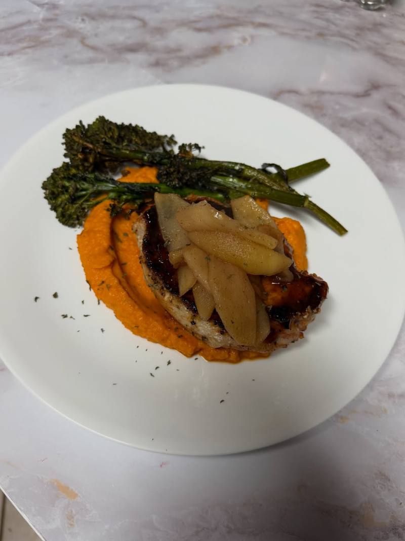 Seared chop topped with sautéed apple slices, served over bright orange purée with charred broccolini on a white plate on a marble countertop.