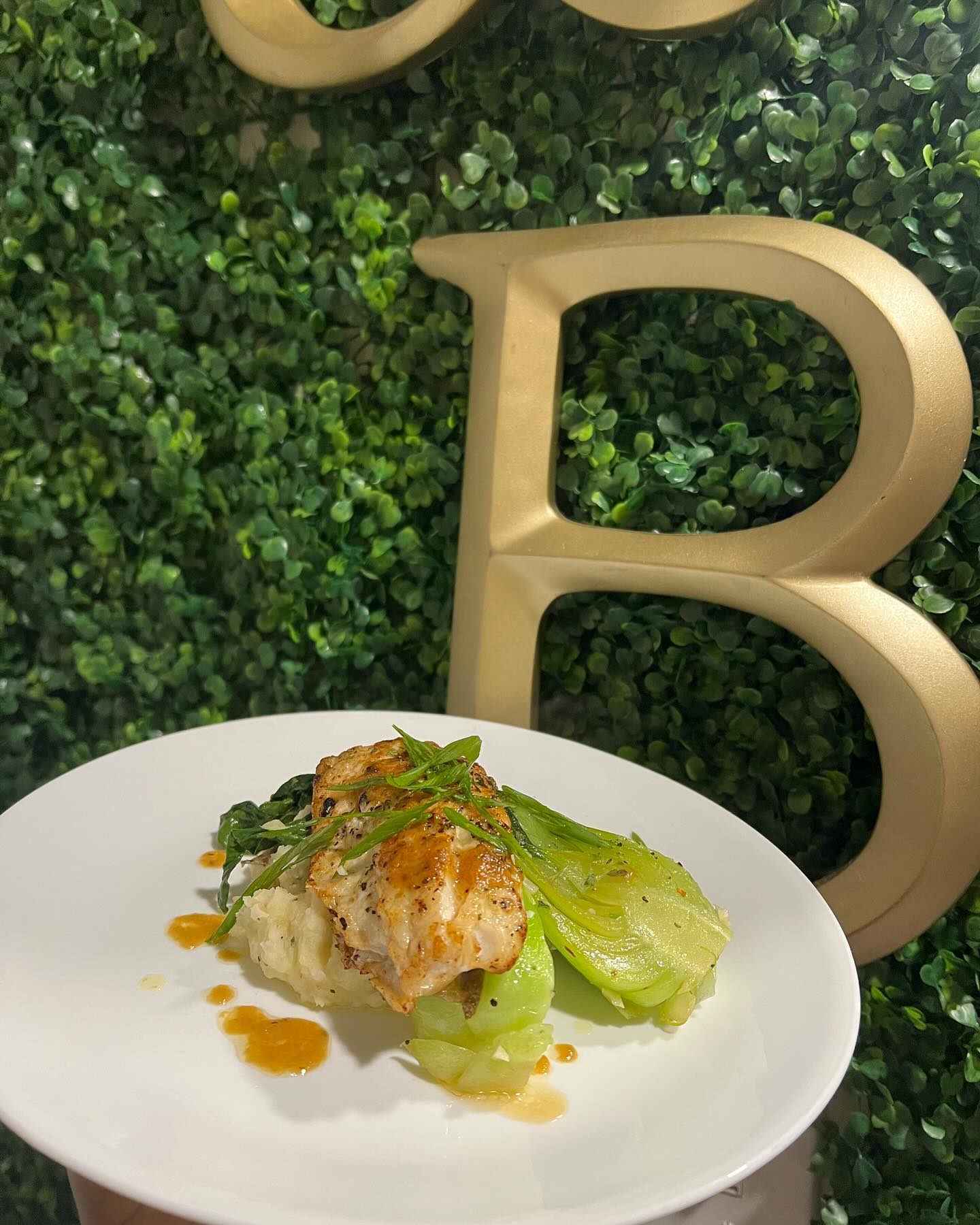 Pan-seared white fish over mashed potatoes and greens with baby bok choy, scallion garnish, and amber sauce on a white plate against a greenery wall with a large gold letter B.
