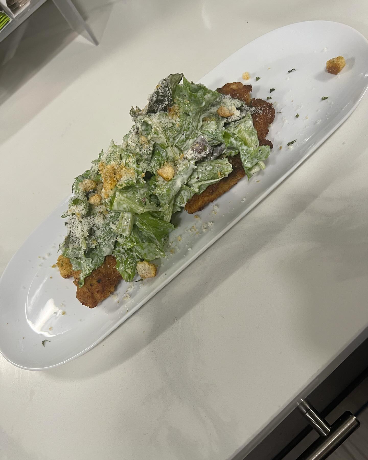 A crispy breaded chicken cutlet topped with Caesar salad, grated Parmesan, and croutons is served on a long white oval platter on a light countertop.