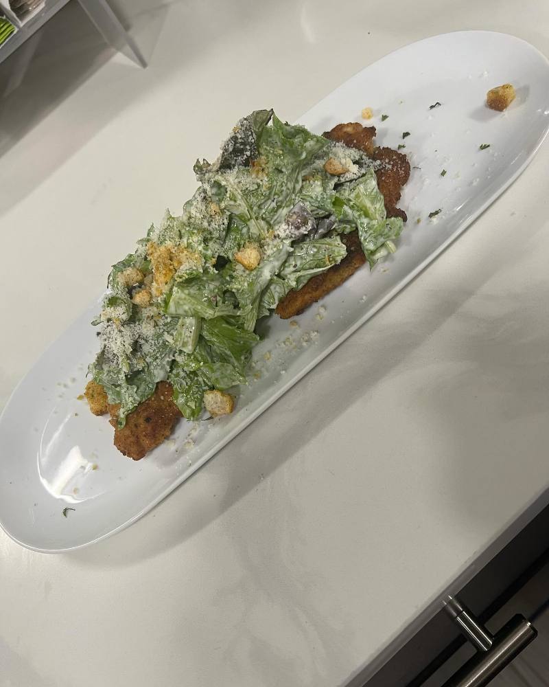 A crispy breaded chicken cutlet topped with Caesar salad, grated Parmesan, and croutons is served on a long white oval platter on a light countertop.