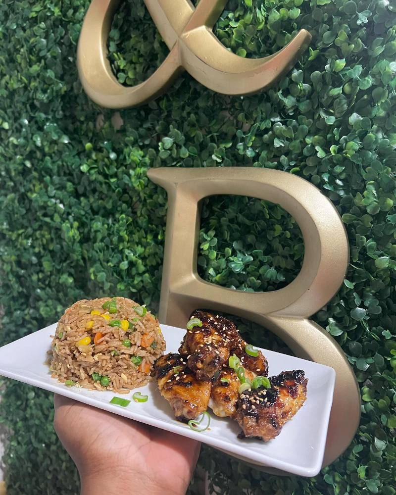 A hand holds a white rectangular plate with a molded mound of vegetable fried rice and glazed chicken wings topped with sesame seeds and green onions against a leafy green wall with gold lettering.