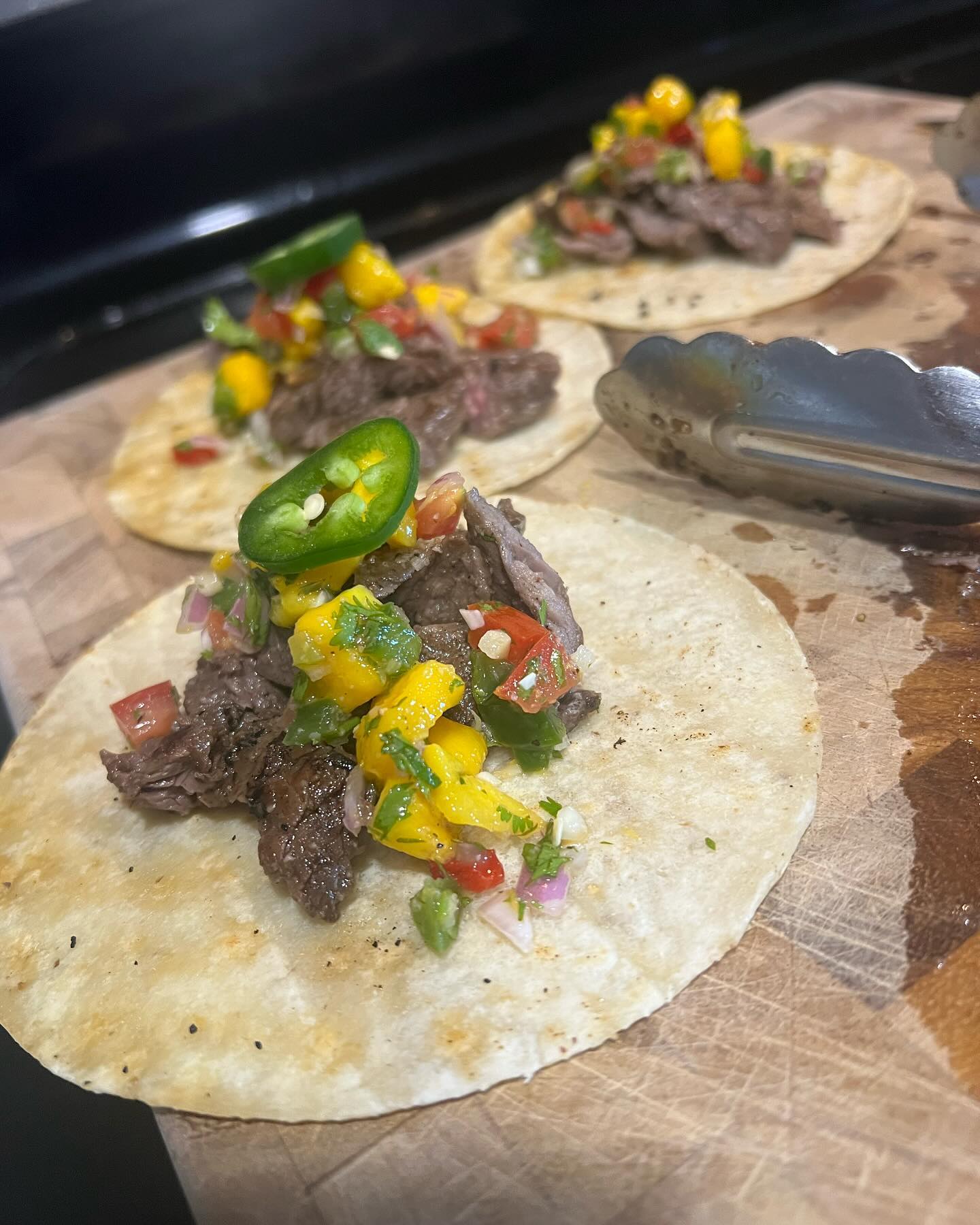 Three steak tacos on corn tortillas topped with mango salsa and jalapeño slices sit on a wooden cutting board beside metal kitchen tongs.
