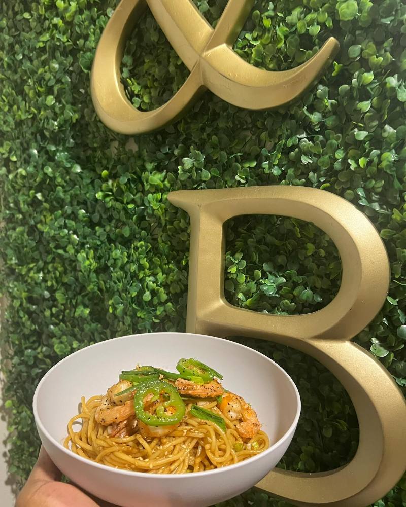 A white bowl of glossy shrimp noodles topped with sliced jalapeños, green onions, and sesame seeds is held in front of a leafy green wall with large gold letters.