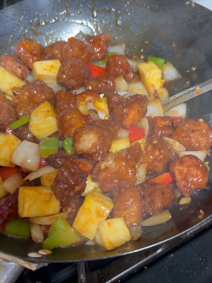 Glossy sweet and sour pork with pineapple chunks, onions, and red and green bell peppers is being stir-fried in a dark wok with a metal spatula on the stovetop.