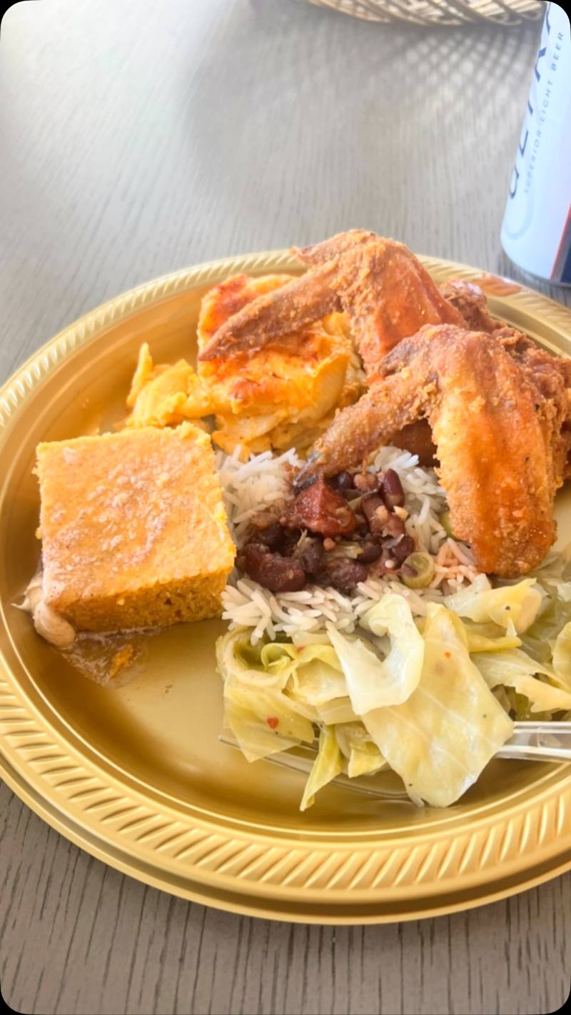 A gold disposable plate on a wooden table holds crispy fried chicken wings, baked macaroni and cheese, cornbread, white rice with red beans, and braised cabbage, with a drink can in the background.