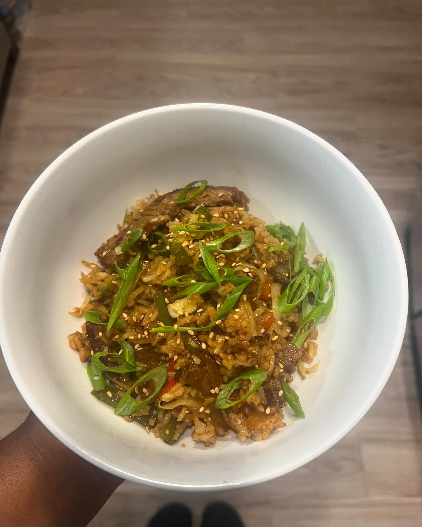 A white bowl of beef fried rice with vegetables, sliced scallions, and sesame seeds is held over a light wood floor.
