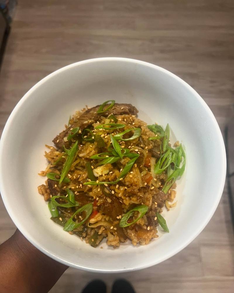 A white bowl of beef fried rice with vegetables, sliced scallions, and sesame seeds is held over a light wood floor.