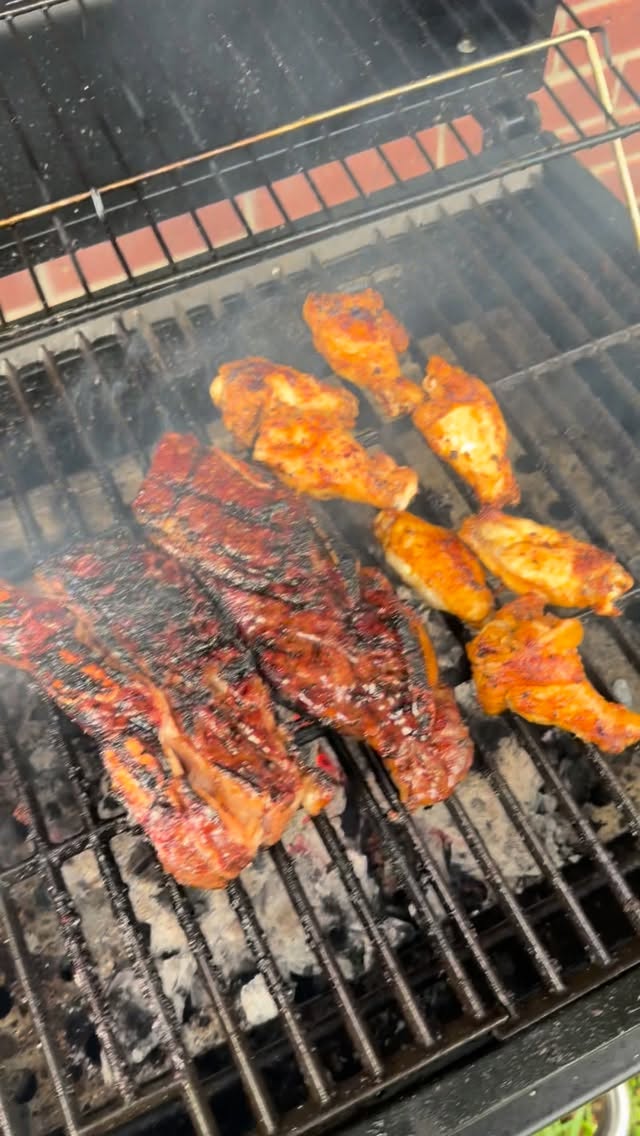 Charred glazed ribs and orange-seasoned chicken pieces cook over smoky charcoal on an outdoor grill.