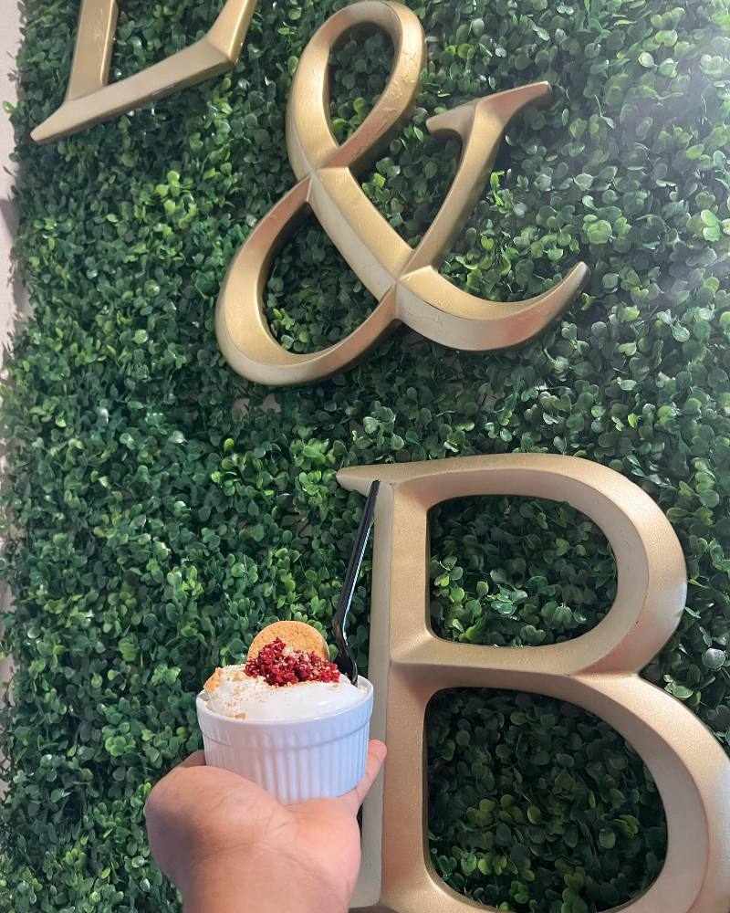 A hand holds a white ribbed ramekin of creamy dessert topped with a vanilla wafer, red velvet crumbles, cookie crumbs, and a black spoon in front of a green hedge wall with gold letters.