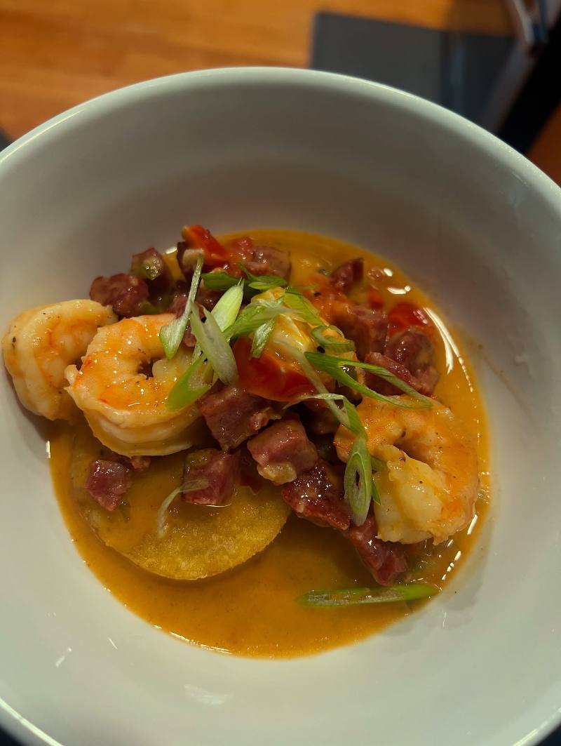 Seared shrimp and diced sausage in orange Creole gravy are plated over a golden grit cake and garnished with sliced green onions in a deep white bowl on a wooden table.