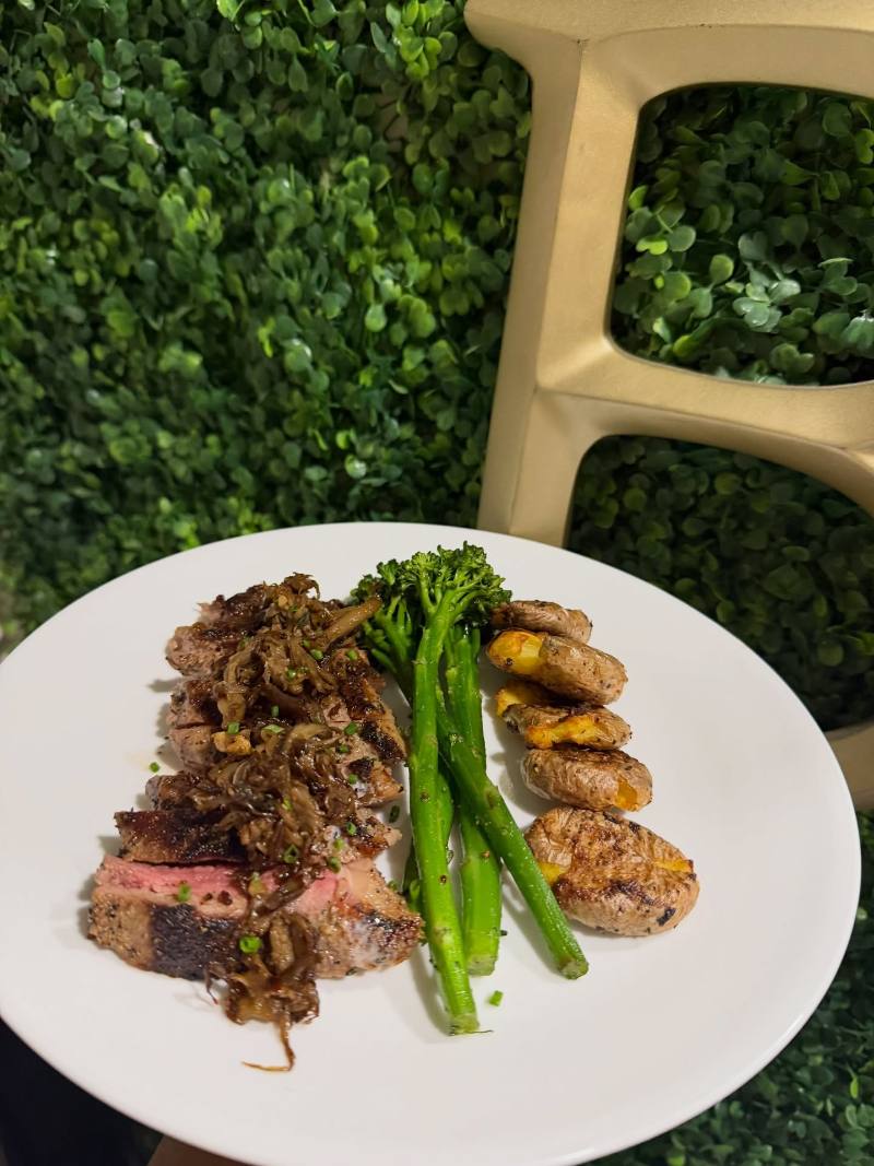 A white plate with sliced steak topped with caramelized onions and chives, broccolini, and crispy smashed potatoes, photographed against a leafy green wall and gold chair.