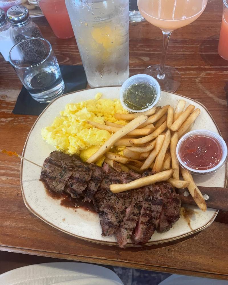 Sliced grilled steak, fluffy scrambled eggs, and French fries are served on a speckled oval plate with cups of chimichurri and ketchup on a wooden table beside drinks.